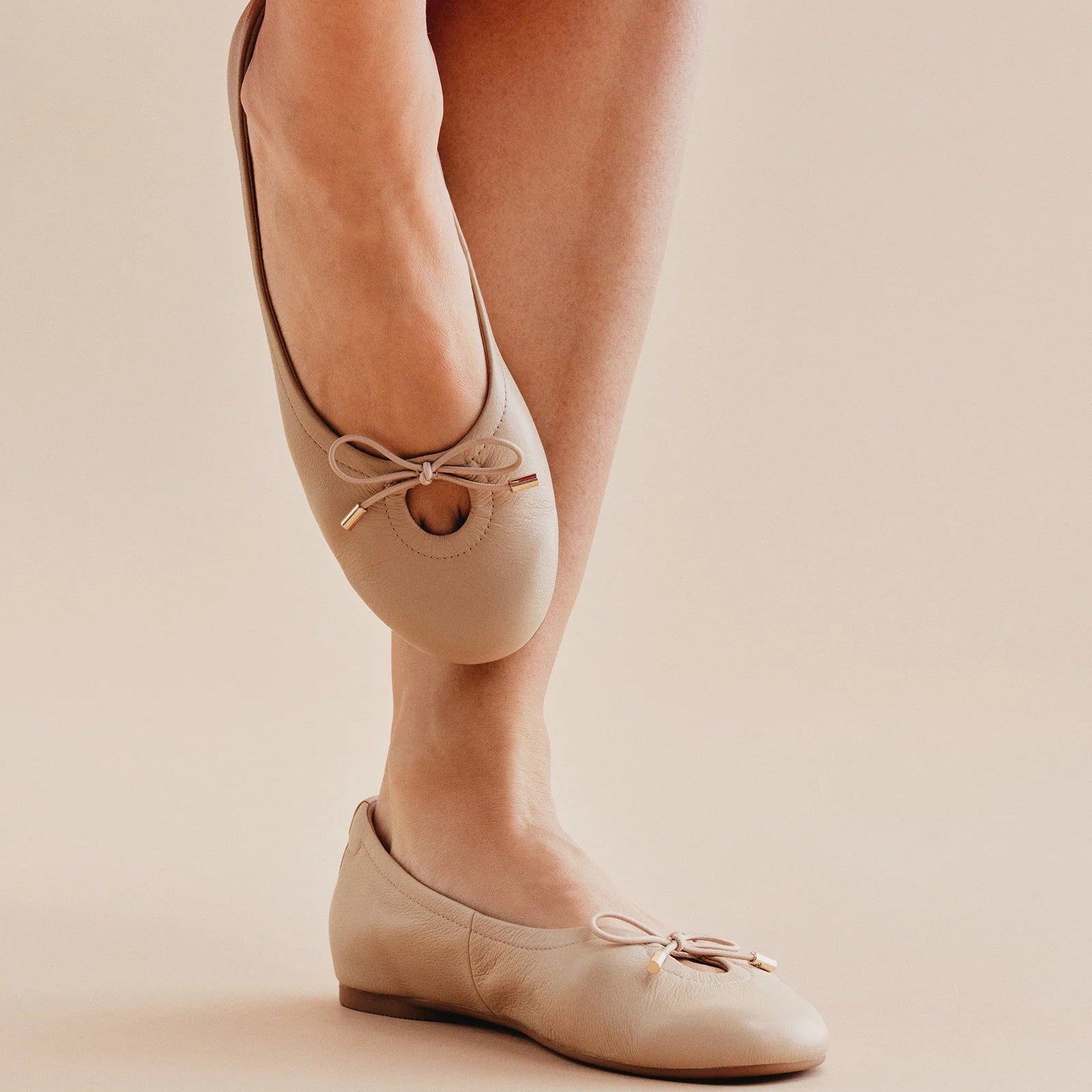 Beige ballet flats with bow detail worn by a person in a red skirt on a beige background