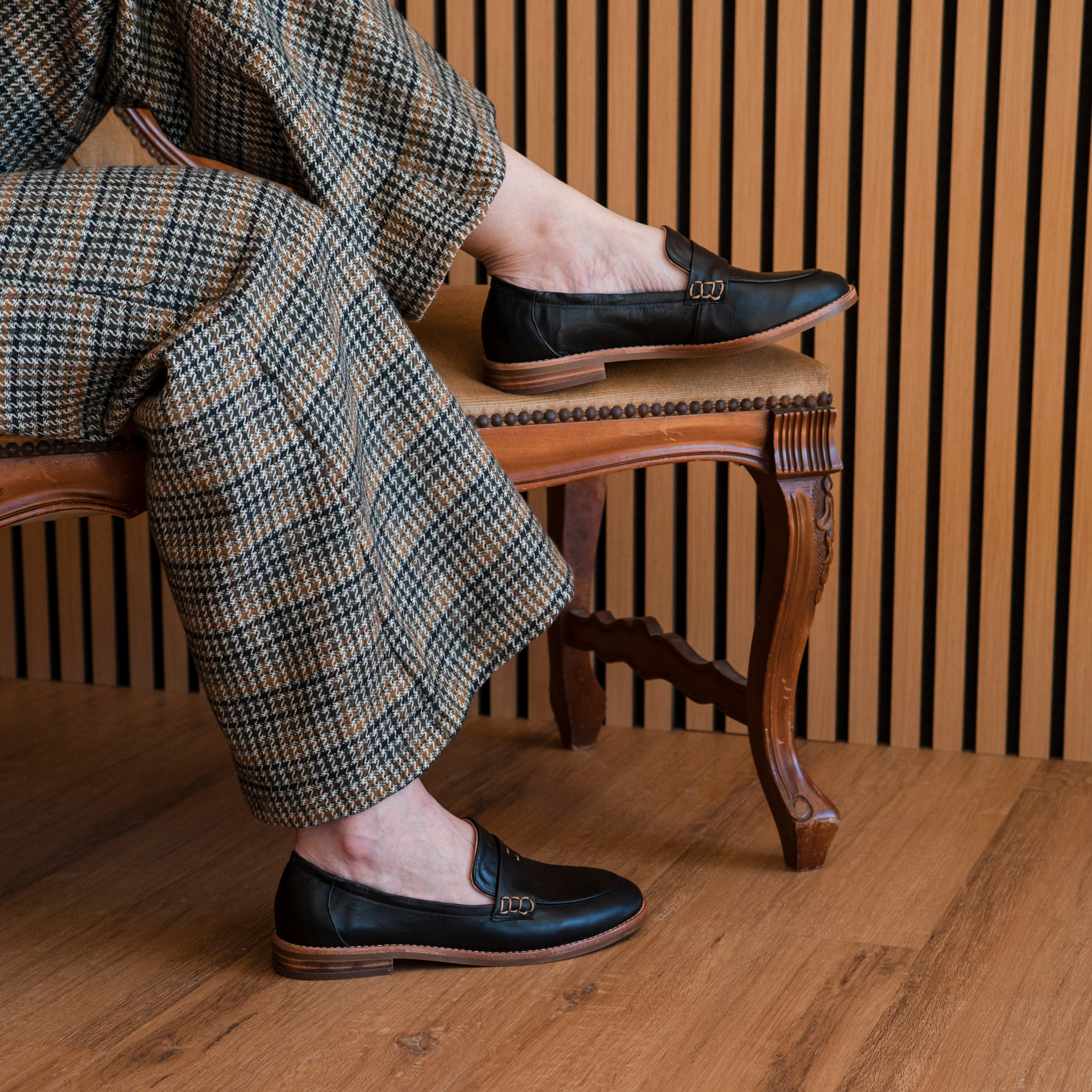 Person wearing checkered pants and black loafers sitting on a wooden chair.