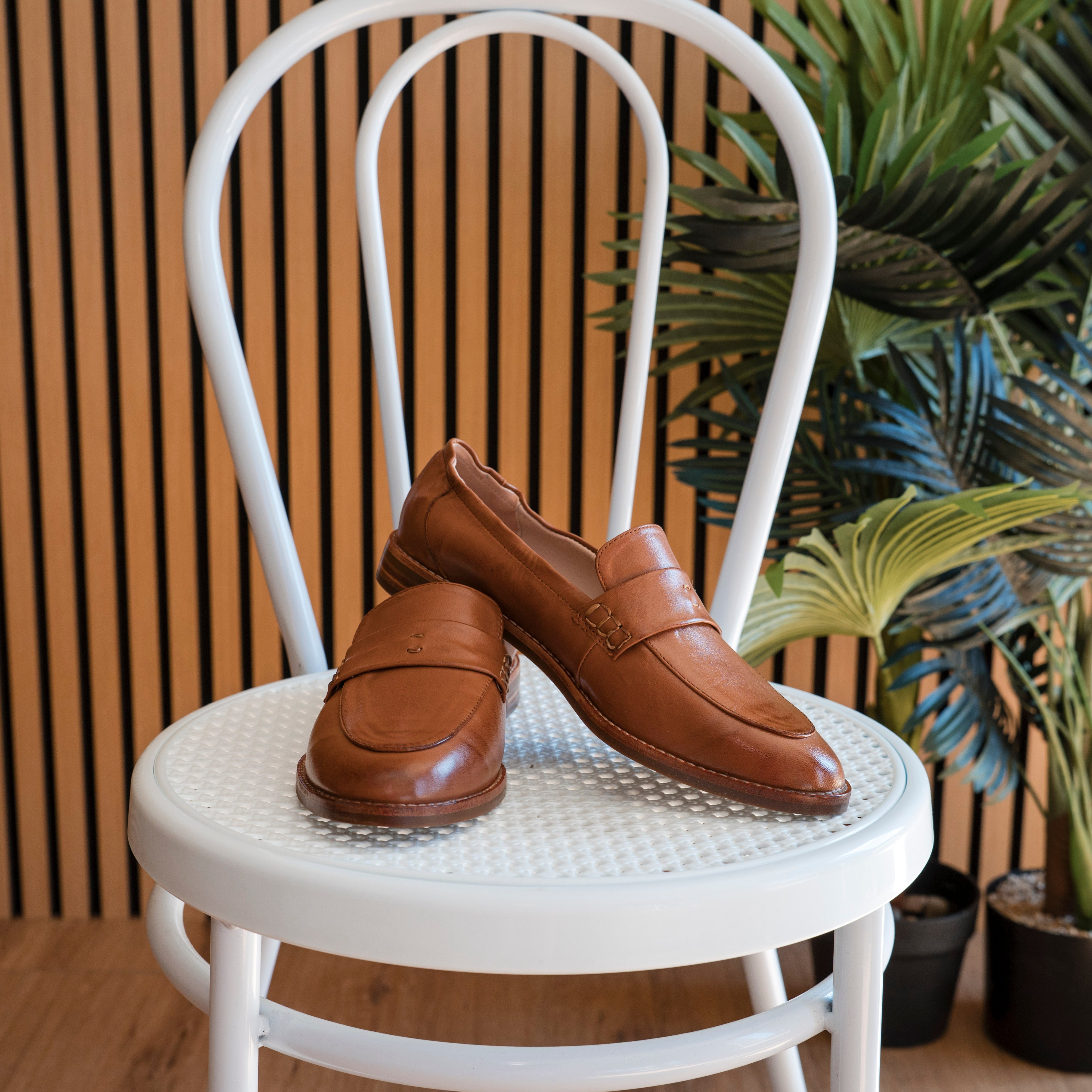 Brown loafers on a white chair with a striped wall and plants in the background