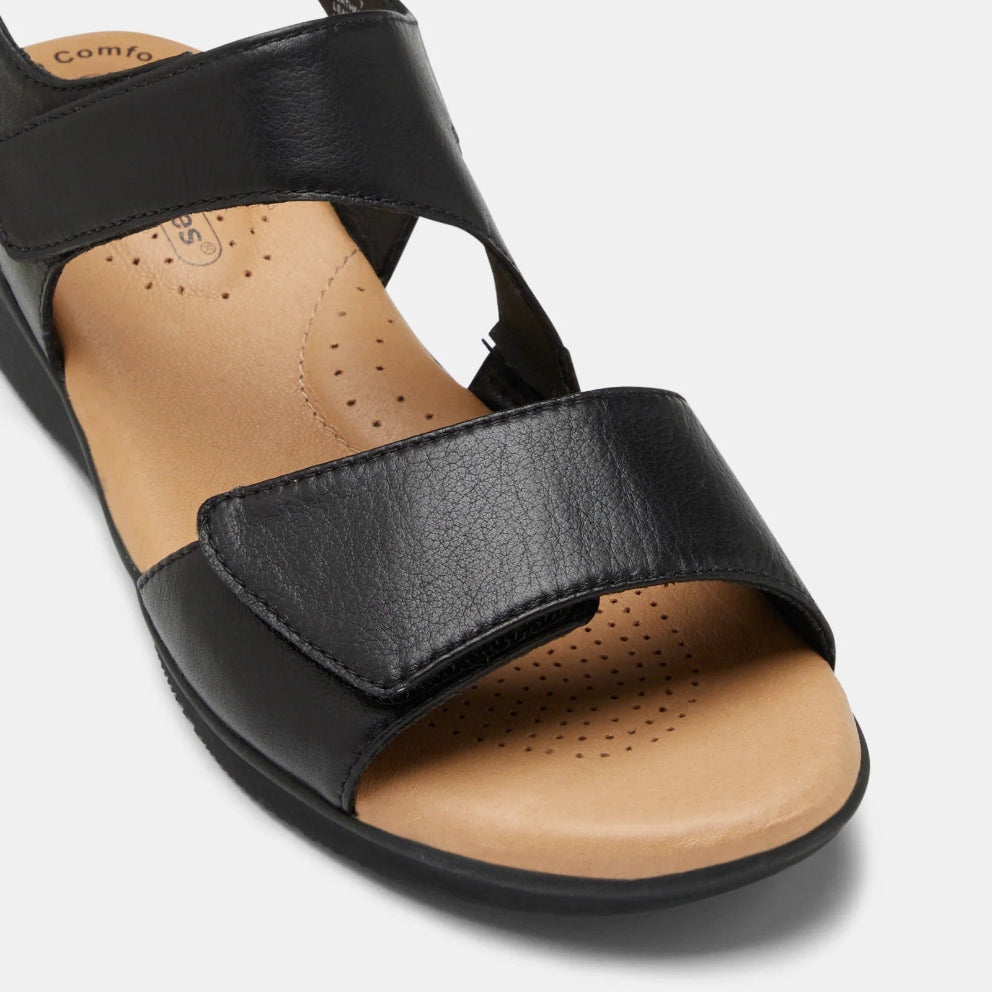 A black leather sandal with a wide adjustable strap and a tan footbed, shown from a high-angle close-up on a light gray background.